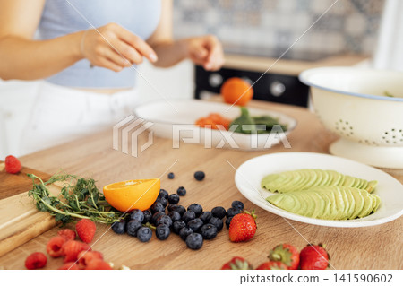 A young girl is preparing a vegan breakfast in the kitchen. Close-up of a plate with avocado, arugula, berries and citrus fruits on a wooden table. A young girl is preparing a vegan breakfast in the kitchen. Close-up of a plate with avocado, arugula, berries and citrus fruits on a wooden table. 141590602