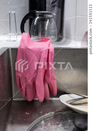 Pink rubber cleaning gloves hanging over a stainless steel kitchen sink beside dishes, showing dishwashing, household chores, hygiene and everyday domestic cleaning routine tasks Pink rubber cleaning gloves hanging over a stainless steel kitchen sink beside dishes, showing dishwashing, household chores, hygiene and everyday domestic cleaning routine tasks 141591513
