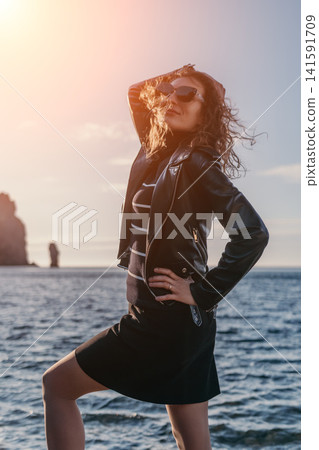 Woman ocean coastline wearing a black leather jacket and sunglasses posing on a rocky shore during a bright sunset Woman ocean coastline wearing a black leather jacket and sunglasses posing on a rocky shore during a bright sunset 141591709