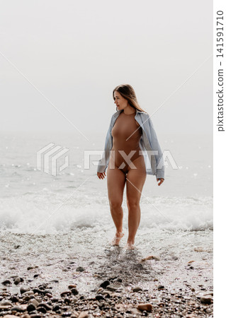 Woman beach ocean walking through gentle waves on a rocky shore wearing a bodysuit and open shirt in serene light Woman beach ocean walking through gentle waves on a rocky shore wearing a bodysuit and open shirt in serene light 141591710