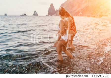Women beach sea two women wading in shallow ocean water on a serene pebble beach at sunset. Women beach sea two women wading in shallow ocean water on a serene pebble beach at sunset. 141591725