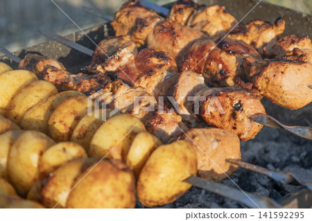 Delicious meat shashlik skewers and golden potatoes roasting on metal skewers over hot charcoal in a grill outdoors. Summer barbecue party food cooking process close-up photography. Delicious meat shashlik skewers and golden potatoes roasting on metal skewers over hot charcoal in a grill outdoors. Summer barbecue party food cooking process close-up photography. 141592295