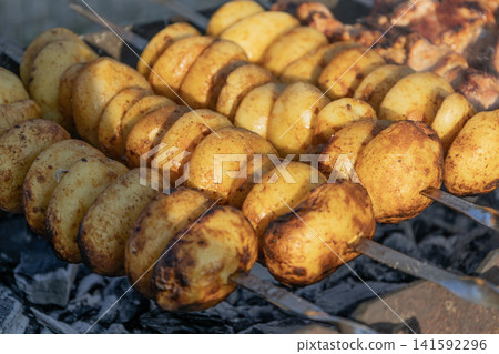 Delicious meat shashlik skewers and golden potatoes roasting on metal skewers over hot charcoal in a grill outdoors. Summer barbecue party food cooking process close-up photography. Delicious meat shashlik skewers and golden potatoes roasting on metal skewers over hot charcoal in a grill outdoors. Summer barbecue party food cooking process close-up photography. 141592296