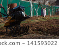 Walk-behind tractor tilling fresh garden soil in a rural homestead, preparing seedbed for spring planting small-scale farming machinery at work in countryside fields. 141593022