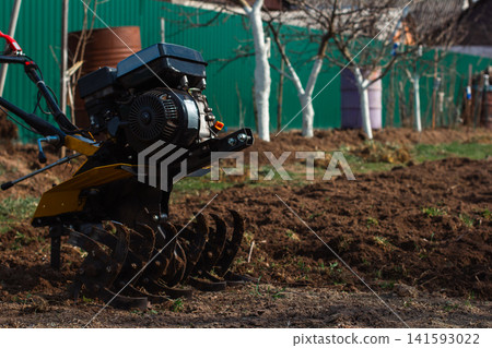 Walk-behind tractor tilling fresh garden soil in a rural homestead, preparing seedbed for spring planting small-scale farming machinery at work in countryside fields. Walk-behind tractor tilling fresh garden soil in a rural homestead, preparing seedbed for spring planting small-scale farming machinery at work in countryside fields. 141593022