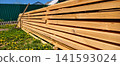Stack of freshly prepared wooden planks sitting on green grass with dandelions. A pitchfork leaning against them. And a green corrugated fence in the background. 141593024