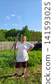 Man smiling and holding a traditional scythe, standing in a sunny backyard with tall green grass, ready for agricultural work and showing dedication to farming and land care. 141593025