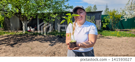Happy woman holding a young green plant in a pot with soil, preparing to plant it in her home garden on a sunny spring day, embracing sustainability and growth. Happy woman holding a young green plant in a pot with soil, preparing to plant it in her home garden on a sunny spring day, embracing sustainability and growth. 141593027