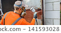 Worker preparing and spray painting an old rusty metal door or gate outdoors with a paint sprayer, wearing protective gear like a respirator mask, gloves, and overalls for renovation and repair. 141593028