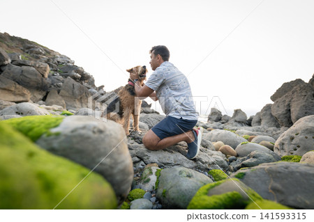 Man kneels on rocks by the sea while interacting with a dog during a sunny day near the coastline Man kneels on rocks by the sea while interacting with a dog during a sunny day near the coastline 141593315