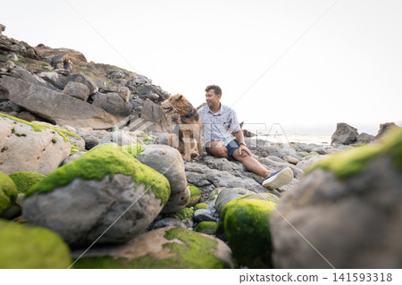 Man sits on rocks by the ocean with his dog during a sunny day at the beach Man sits on rocks by the ocean with his dog during a sunny day at the beach 141593318