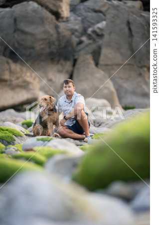 Man and dog sit on rocky shore with green moss during late afternoon Man and dog sit on rocky shore with green moss during late afternoon 141593325