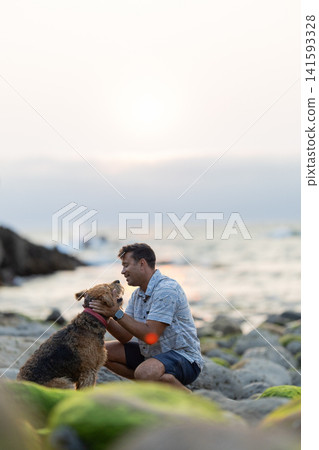 Man interacts with dog at the beach during sunset on a rocky shore near the water Man interacts with dog at the beach during sunset on a rocky shore near the water 141593328