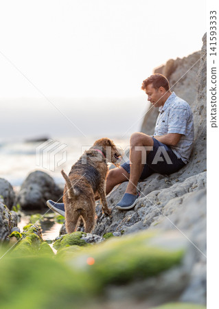 Man sits on rocks by the sea with dog during sunset near the shore 141593333
