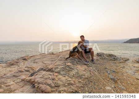 Man plays with a dog on a rocky shore while the sun sets over the ocean Man plays with a dog on a rocky shore while the sun sets over the ocean 141593348