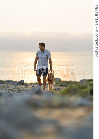 Man walks a dog along the rocky shore during sunset near the water's edge Man walks a dog along the rocky shore during sunset near the water's edge 141593465