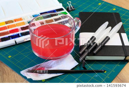 Artistic supplies for watercolor painting and hobby as  a watercolor paint set, a brush pens, sketchbook and water in a glass cup on a mat. 141593674