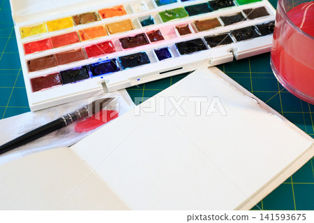 Artistic tools for watercolor painting and hobby as open sketchbook, a watercolor paint set, a brush, and water in a glass cup 141593675