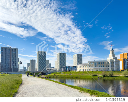 Modern urban landscape featuring tall residential buildings alongside a river, with a park area in the foreground. The scene is set against a clear blue sky with scattered clouds Modern urban landscape featuring tall residential buildings alongside a river, with a park area in the foreground. The scene is set against a clear blue sky with scattered clouds 141593819