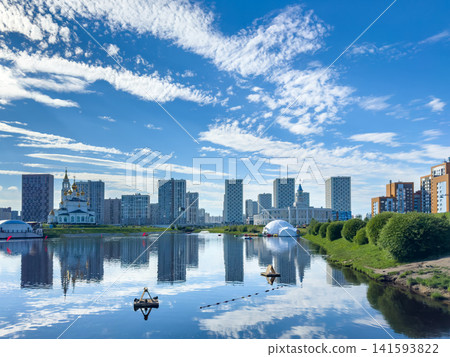Urban skyline featuring modern high-rise buildings along a river, with reflections visible on the water surface. Lush greenery and a distant church add to the scenic view of the city Urban skyline featuring modern high-rise buildings along a river, with reflections visible on the water surface. Lush greenery and a distant church add to the scenic view of the city 141593822