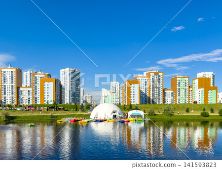 Modern residential buildings line the shore of a tranquil lake, featuring recreational boats and a white dome structure, under a clear blue sky with a few clouds visible 141593823