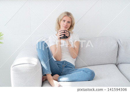 Woman enjoying cozy afternoon. Relaxed lady with coffee relaxes. Serene woman on sofa with coffee. Casual woman relaxing indoors with warm beverage and peaceful ambiance Woman enjoying cozy afternoon. Relaxed lady with coffee relaxes. Serene woman on sofa with coffee. Casual woman relaxing indoors with warm beverage and peaceful ambiance 141594306