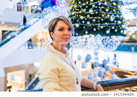 Woman standing in festive shopping mall near Christmas tree. Shopper enjoying holiday decorations and atmosphere. Winter season sale and celebration concept in retail center interior. Woman standing in festive shopping mall near Christmas tree. Shopper enjoying holiday decorations and atmosphere. Winter season sale and celebration concept in retail center interior. 141594484