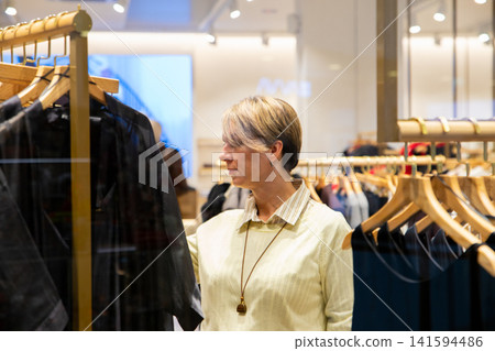 Woman browsing clothes in store. Customer looking at garment on hanger in boutique. Shopping experience, fashion retail concept. Consumer searching for apparel in clothing store interior. 141594486