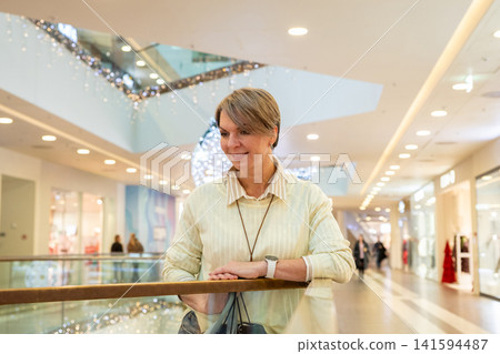 Woman standing in shopping mall with luxury store interior. Mature lady holding shopping bag during seasonal sale. Retail lifestyle concept with consumer activity in urban commercial center. Woman standing in shopping mall with luxury store interior. Mature lady holding shopping bag during seasonal sale. Retail lifestyle concept with consumer activity in urban commercial center. 141594487