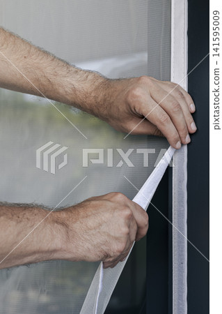 Hands adjusting a mosquito screen on a window frame, illustrating seasonal home preparations and preventive care in urban setting. Hands adjusting a mosquito screen on a window frame, illustrating seasonal home preparations and preventive care in urban setting. 141595009