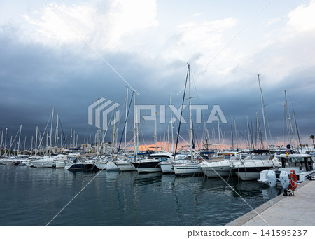 Numerous yachts and sailboats moored at a marina pier under a cloudy sky during twilight Numerous yachts and sailboats moored at a marina pier under a cloudy sky during twilight 141595237