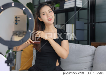 Young Asian woman showing hair tutorial in front of phone camera and ring light. Young Asian woman showing hair tutorial in front of phone camera and ring light. 141595588