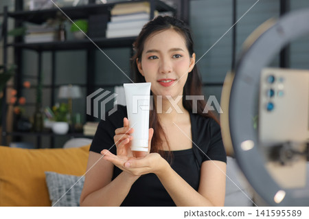 Asian woman filming beauty vlog tutorial with ring light, reviewing cosmetics cream moisturizer or lotion. Asian woman filming beauty vlog tutorial with ring light, reviewing cosmetics cream moisturizer or lotion. 141595589
