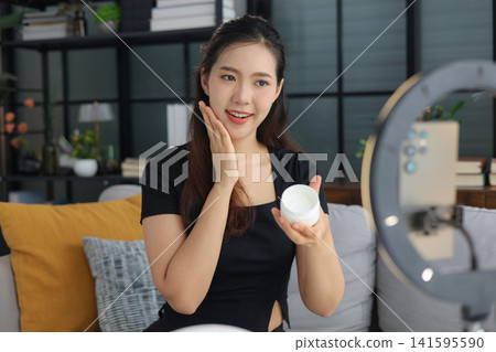 Asian woman filming beauty vlog tutorial with ring light, reviewing cosmetics cream moisturizer or lotion. Asian woman filming beauty vlog tutorial with ring light, reviewing cosmetics cream moisturizer or lotion. 141595590