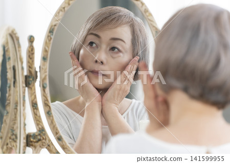 一位老年婦女在鏡子前進行護膚步驟。 一位老年婦女在鏡子前進行護膚步驟。 141599955
