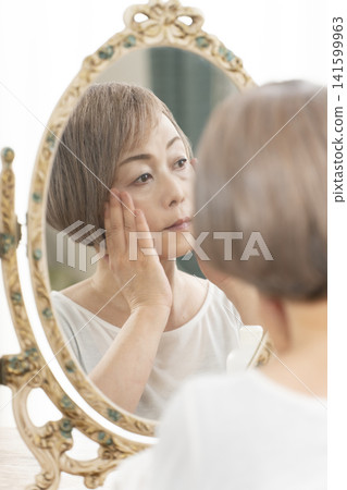 一位老年婦女在鏡子前進行護膚步驟。 一位老年婦女在鏡子前進行護膚步驟。 141599963