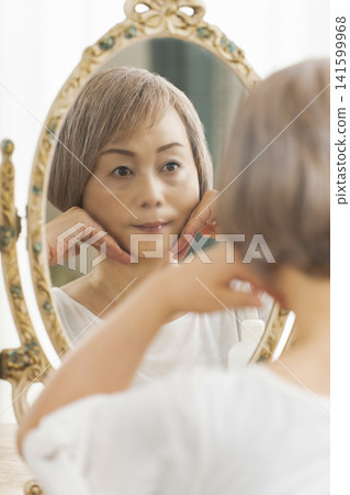 一位老年婦女在鏡子前進行護膚步驟。 一位老年婦女在鏡子前進行護膚步驟。 141599968