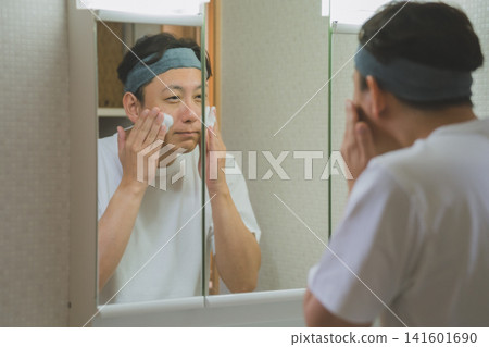 A man in his 40s, wearing a headband, washes his face with foam in front of a mirror. A man in his 40s, wearing a headband, washes his face with foam in front of a mirror. 141601690