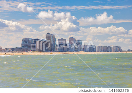Ostend, a coastal city in Belgium, Europe. Coastal cityscape view from sea under blue sky with white clouds, beach with distant figures and modern buildings on horizon Ostend, a coastal city in Belgium, Europe. Coastal cityscape view from sea under blue sky with white clouds, beach with distant figures and modern buildings on horizon 141602276