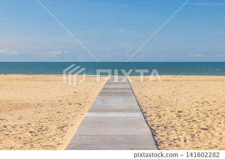 Wooden boardwalk path leading to the vast blue sea with golden sand under a clear sky on a sunny day at the beach. Ostend, a coastal city in Belgium, Europe. Wooden boardwalk path leading to the vast blue sea with golden sand under a clear sky on a sunny day at the beach. Ostend, a coastal city in Belgium, Europe. 141602282