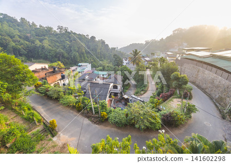 Panoramic high-angle view of a serene hillside village nestled in lush green mountains under soft morning sunlight, showcasing a peaceful tropical landscape. Kandy town in Sri Lanka. Panoramic high-angle view of a serene hillside village nestled in lush green mountains under soft morning sunlight, showcasing a peaceful tropical landscape. Kandy town in Sri Lanka. 141602298