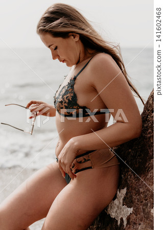 Woman beach swimwear leaning against a coastal rock while holding sunglasses during a serene summer day at the ocean Woman beach swimwear leaning against a coastal rock while holding sunglasses during a serene summer day at the ocean 141602468