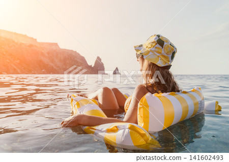 Woman, Beach, Float - Woman relaxing on a yellow and white striped inflatable float in the ocean with cliffs in the distance. 141602493