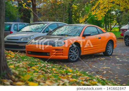 Autumn. Orange modified sports coupe with black hood parked beside other vehicles on a tree-lined street covered with fallen leaves Autumn. Orange modified sports coupe with black hood parked beside other vehicles on a tree-lined street covered with fallen leaves 141602847