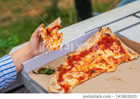 Close-up of child holding and eating slice of freshly baked homemade cheese pizza prepared at master class beside an open cardboard box. Casual outdoor meal after pizza making activity Close-up of child holding and eating slice of freshly baked homemade cheese pizza prepared at master class beside an open cardboard box. Casual outdoor meal after pizza making activity 141603820