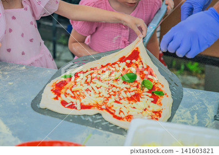 Children Helping Arrange Homemade Pizza on Baking Peel. Children and adult carefully lift and arrange uncooked pizza dough with cheese, tomato sauce and basil on baking peel. Cooking workshop Children Helping Arrange Homemade Pizza on Baking Peel. Children and adult carefully lift and arrange uncooked pizza dough with cheese, tomato sauce and basil on baking peel. Cooking workshop 141603821