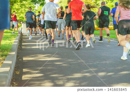 Participants run through park, Become a Hero obstacle race in Burgas. Adult participants run along a park pathway during the Become a Hero sport obstacle race. Rear view Participants run through park, Become a Hero obstacle race in Burgas. Adult participants run along a park pathway during the Become a Hero sport obstacle race. Rear view 141603845
