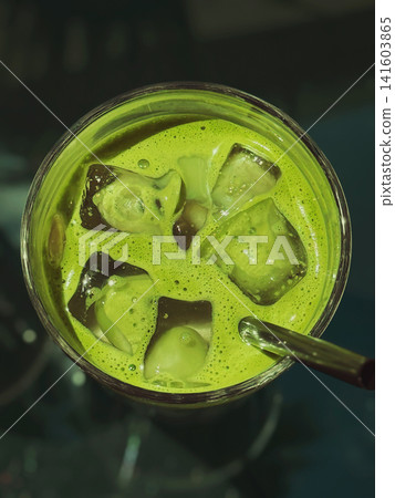Close-up top view of vivid green iced matcha tea in clear glass with large melting ice cubes and drinking straw. Refreshing cold beverage on dark cafe table Close-up top view of vivid green iced matcha tea in clear glass with large melting ice cubes and drinking straw. Refreshing cold beverage on dark cafe table 141603865