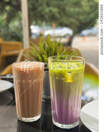 Chocolate milk drink and colorful layered iced matcha latte served in tall glasses on an outdoor cafe table. Modern summer beverage composition with ice, plant and blurred terrace Chocolate milk drink and colorful layered iced matcha latte served in tall glasses on an outdoor cafe table. Modern summer beverage composition with ice, plant and blurred terrace 141603866