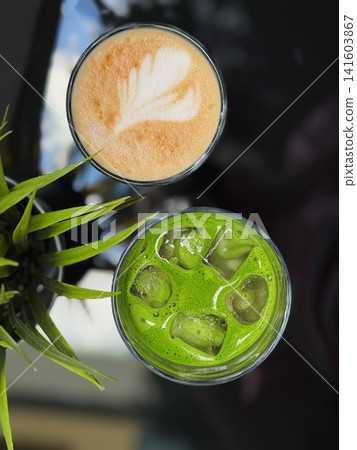 Top view of hot coffee with milk foam and bright green iced matcha drink on reflective cafe table. Modern beverage composition with plant leaves and dark copy space Top view of hot coffee with milk foam and bright green iced matcha drink on reflective cafe table. Modern beverage composition with plant leaves and dark copy space 141603867
